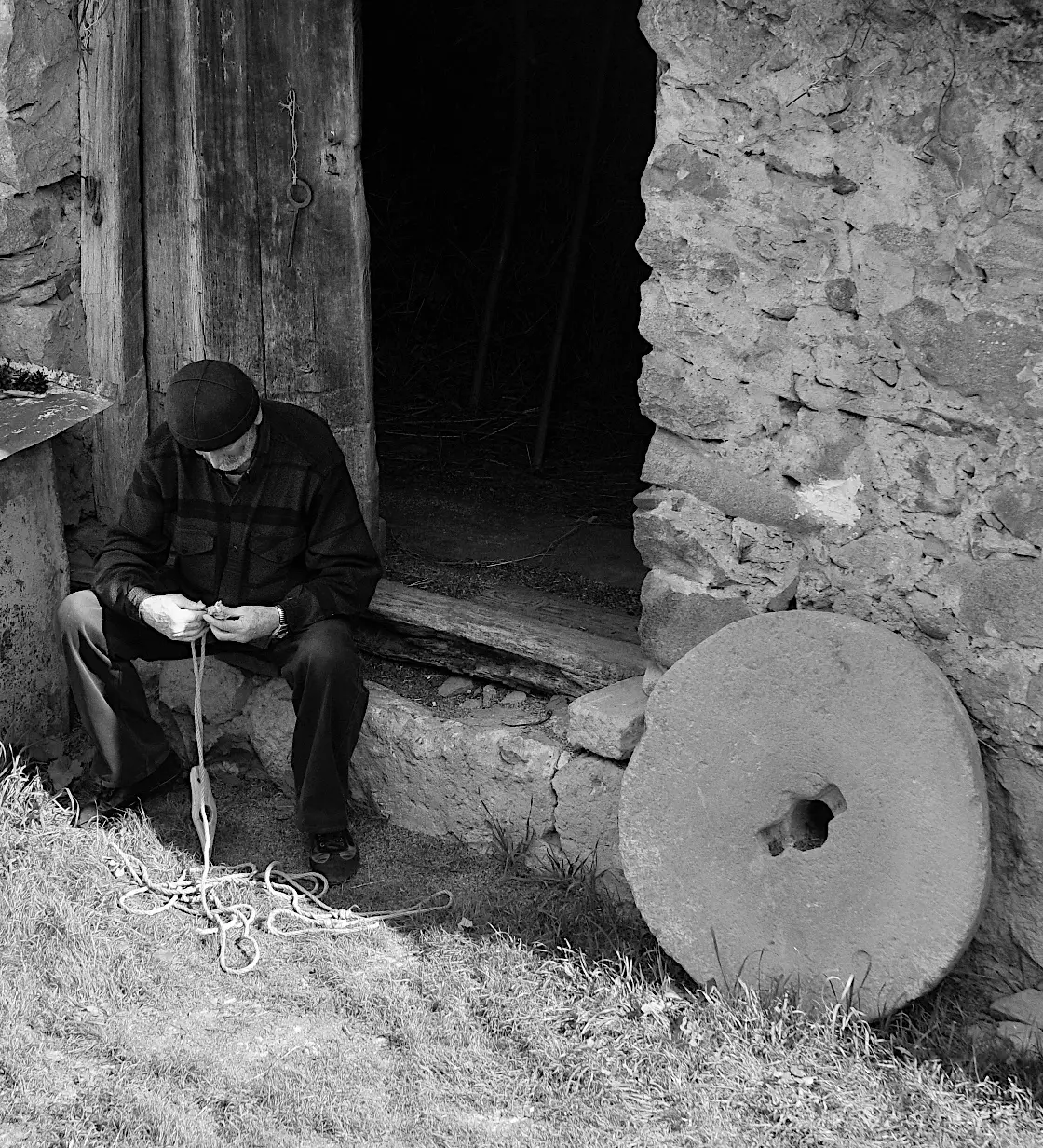 BW photo of local villager man sitting in front of stone house, wearing traditional Svani hat